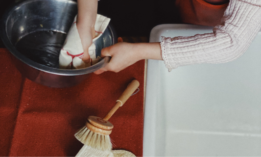 Petite fille concentrée participant à une activité cuisine pour tout-petits, apprenant à laver la vaisselle dans un évier, avec des ustensiles et du savon adaptés à sa taille.